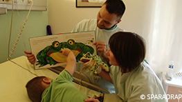 Pendant une injection intra musculaire, un petit garçon est distrait par son papa qui lui lit un livre avec une grenouille en relief, l'auxiliaire de puériculture joue avec une marionnette à main en forme de grenouille.